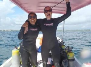 two female scuba divers in wetsuits, smiling, standing on a boat